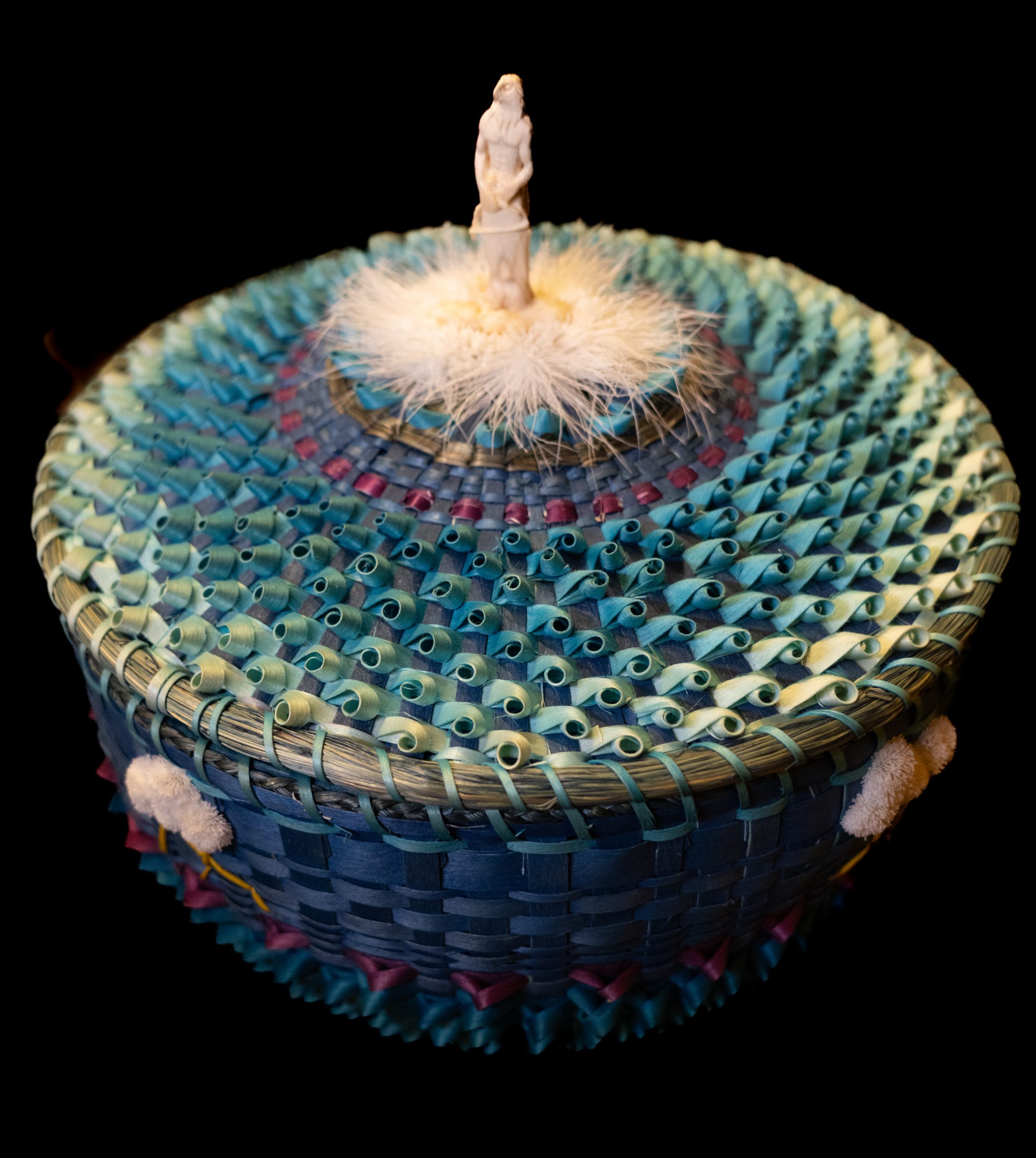 Ohiyo Art Market Winners-19 A basket made in blue and purple and yellow hues. The lid has some white fur in center and a tiny sculpture of a man with an Eagle's head is placed on top.