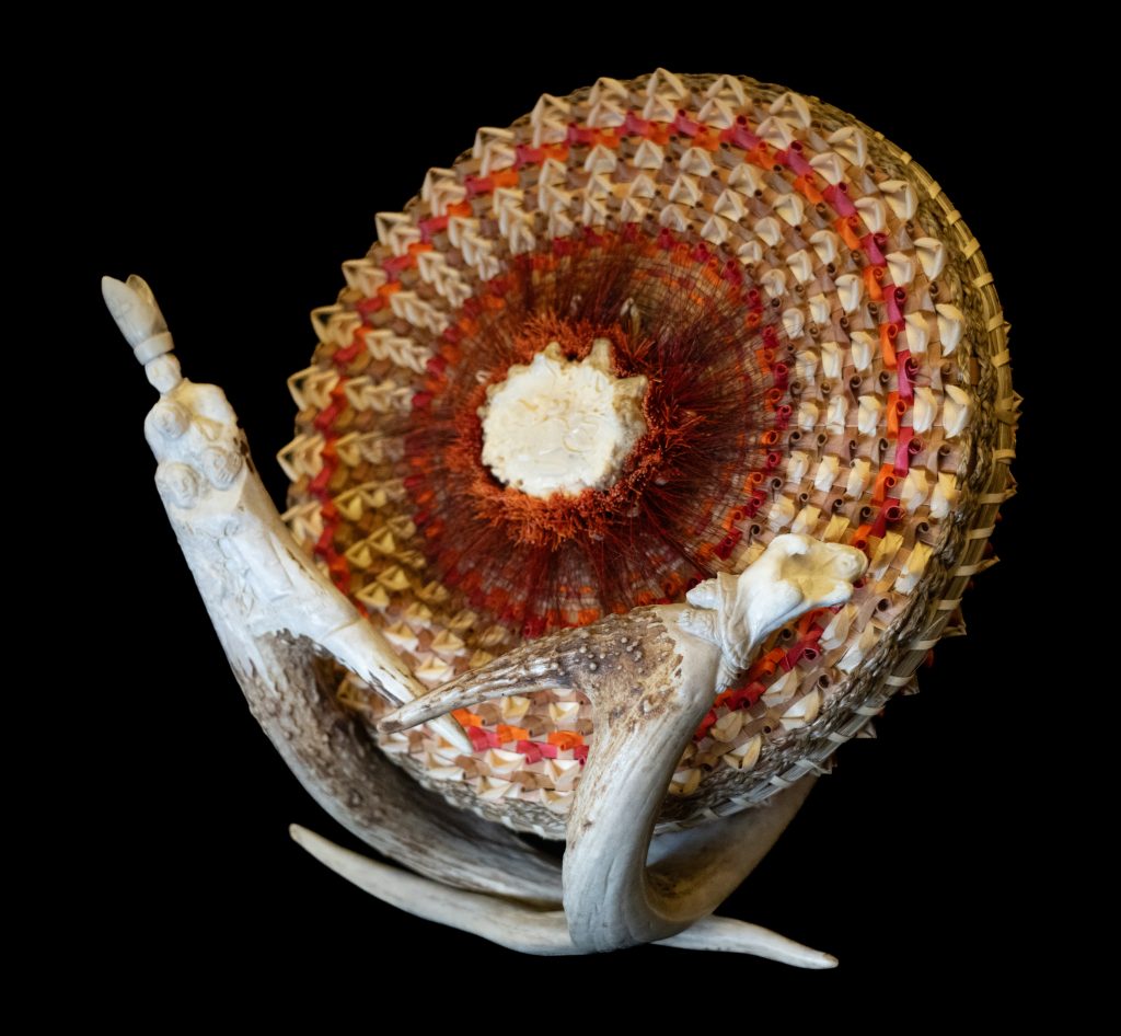 Basket weaved in orange and tan hues. It is mounted on a set of carved antlers. The antlers are carved into a Pope and schoolchildren.