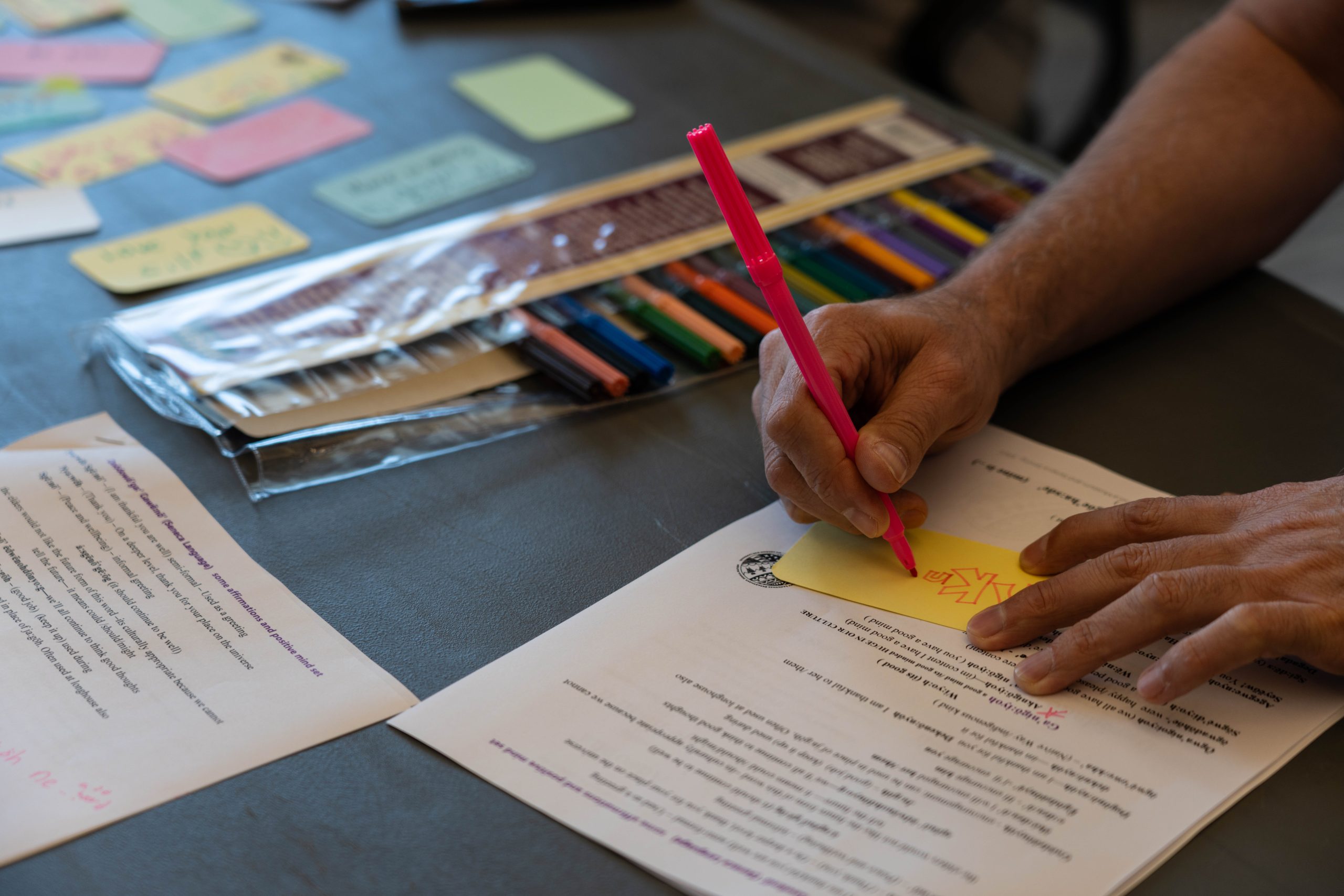 Cropped image of someone actively creating an affirmation card. Photo only shows the person's arms/ They hold a red pen and are writing on a yellow card. There is a reference sheet of words in the Seneca language under the small yellow card.