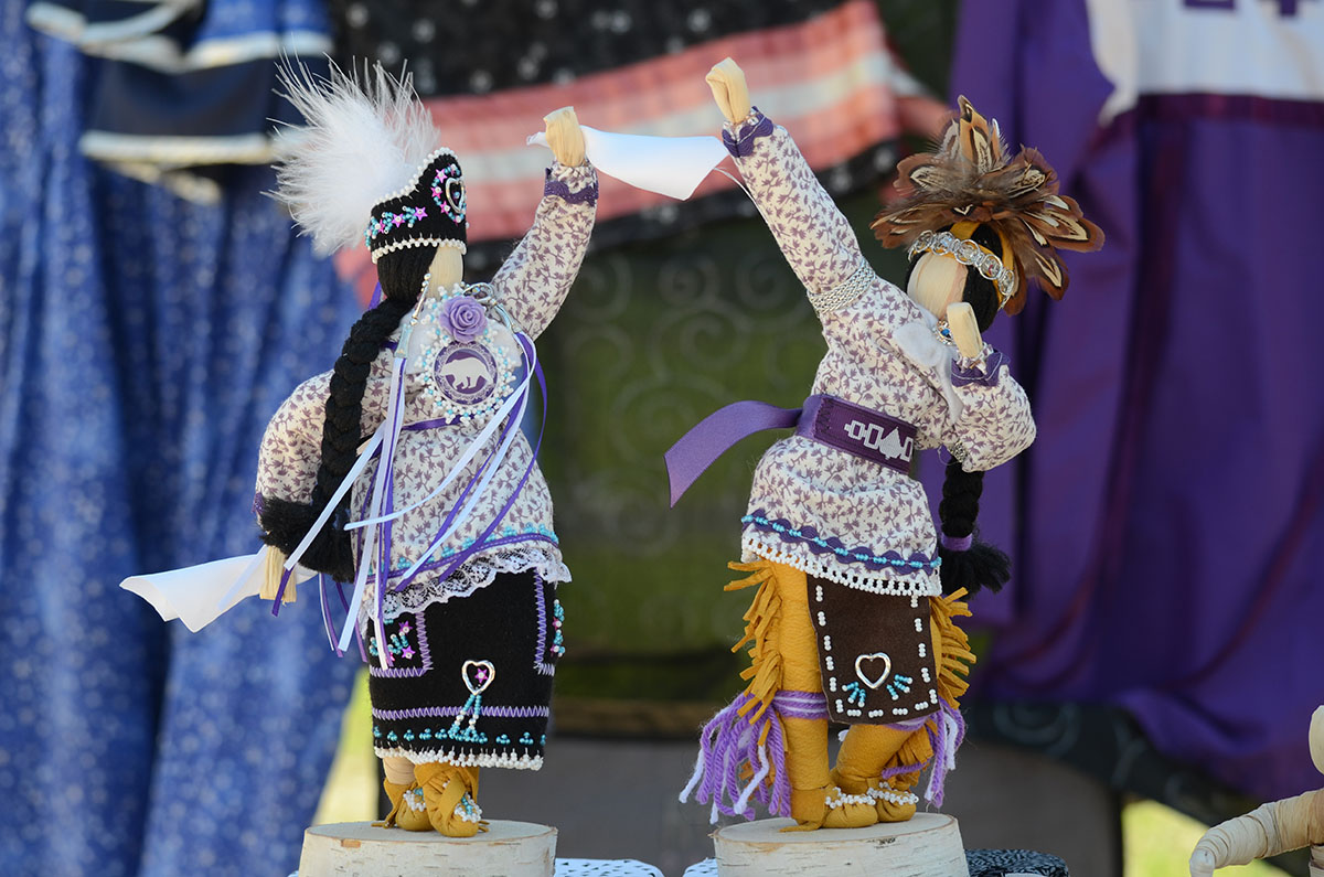 Two cornhusk dolls posed like performing smokedance. The dolls are dressed in traditional Smoke Dance regalia in Haudenosaunee purple.