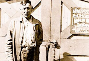 A vintage photo of a man in a jacket and slacks standing in front of the wooden doors of a railroad station building.