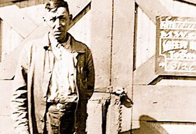 A vintage photo of a man in a jacket and slacks standing in front of the wooden doors of a railroad station building.